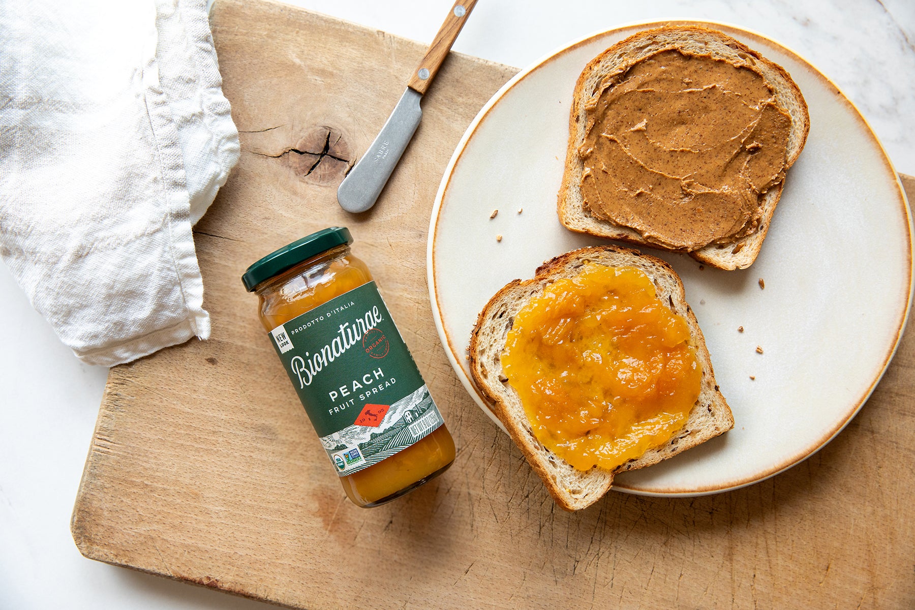 Toast topped with peach fruit spread beside a jar of Bionaturae organic peach fruit spread on a wooden cutting board