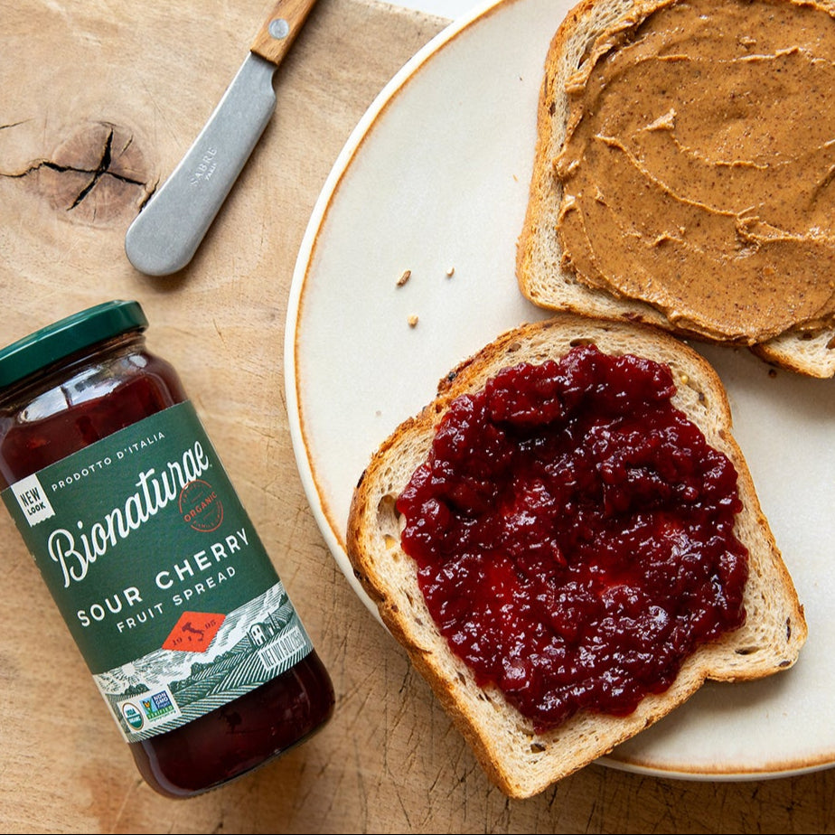 Slice of toast spread with organic sour cherry fruit spread beside an open Bionaturae jar on a wooden table