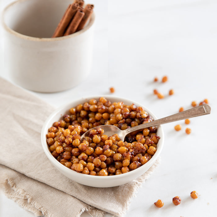 Bowl of organic cinnamon-sugar coated roasted chickpeas with spoon on beige napkin and cinnamon sticks in the background