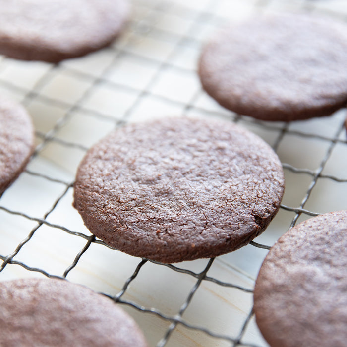 Close-up of an Einkorn chocolate sandwich cookie on a wire cooling rack showing textured cocoa surface with other cookies blurred behind