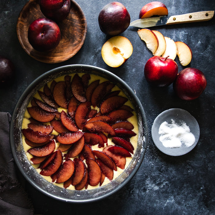 Einkorn plum cake in round pan topped with plum slices on dark countertop with whole plums and a small bowl of sugar