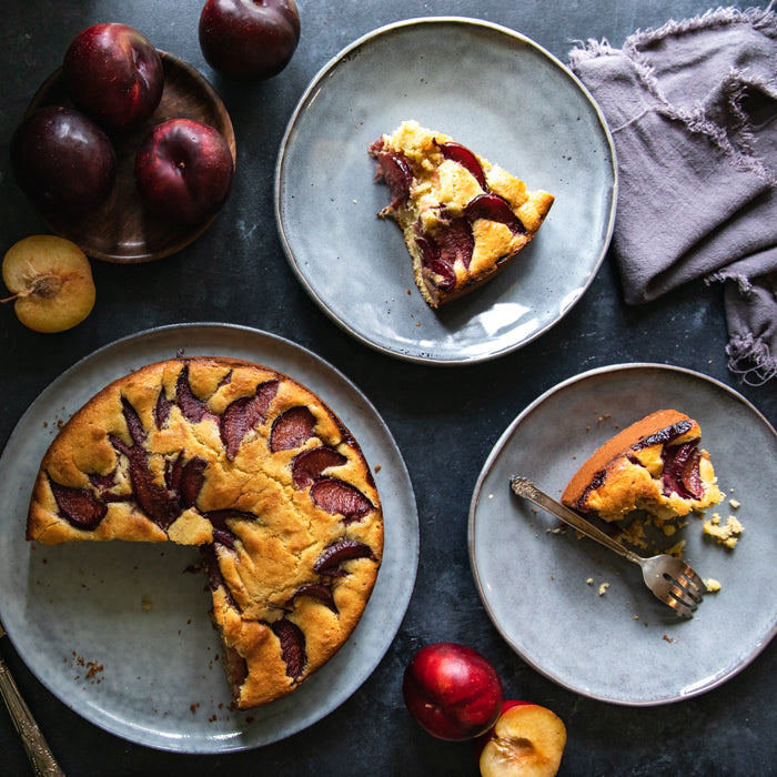 Einkorn plum coffee cake sliced on ceramic plates with fresh plums on a dark tabletop