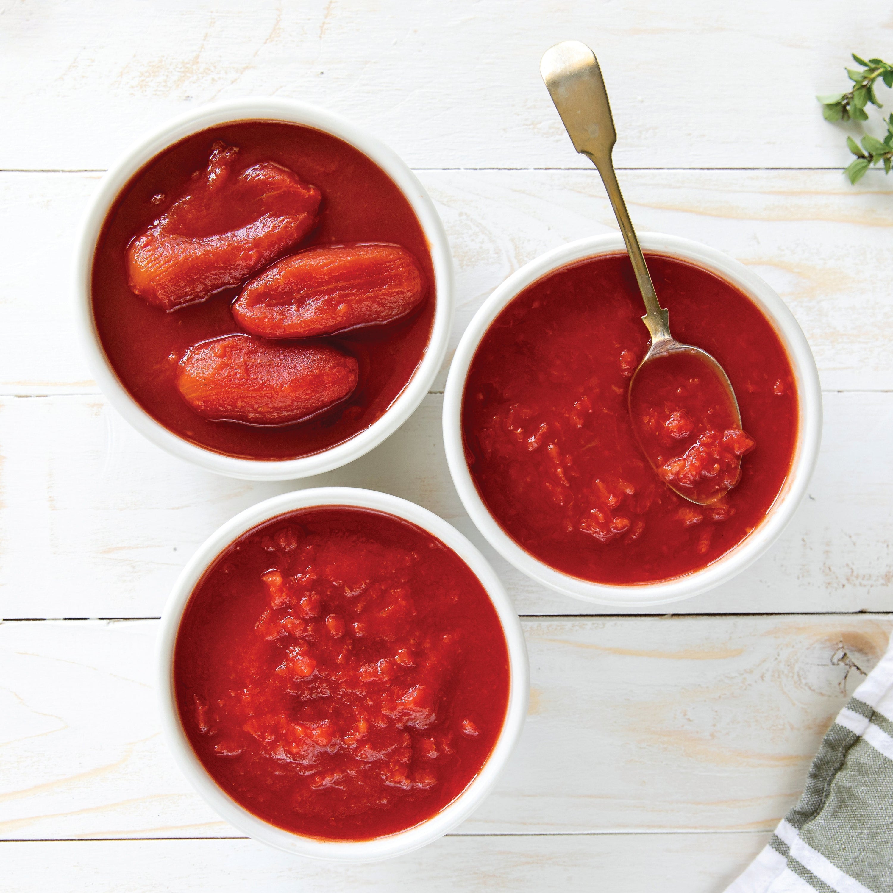 Three bowls of organic Italian crushed tomatoes showing chunky and smooth textures on a white table with a serving spoon