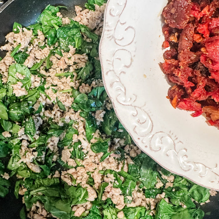 Ground sausage and wilted spinach sautéing in a skillet with a bowl of sun-dried tomatoes at the pan edge