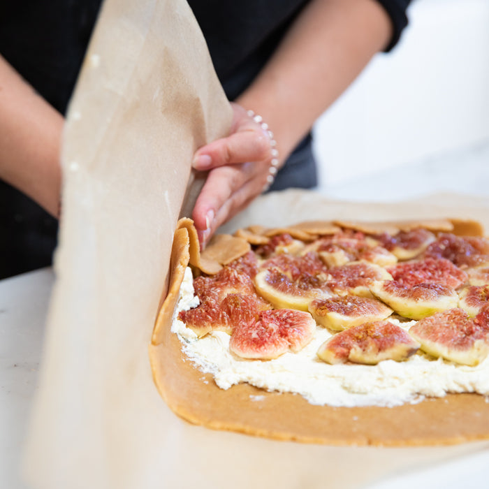 Hands folding parchment over a rustic einkorn galette layered with sliced figs and goat cheese on a marble countertop