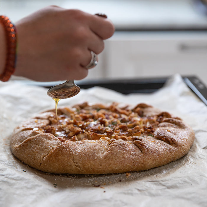 Fig and goat cheese einkorn galette topped with chopped nuts being drizzled with honey on parchment in a home kitchen