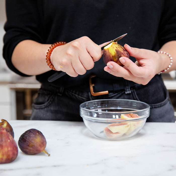 Person peeling a fresh fig into a glass bowl on a marble countertop, whole figs nearby