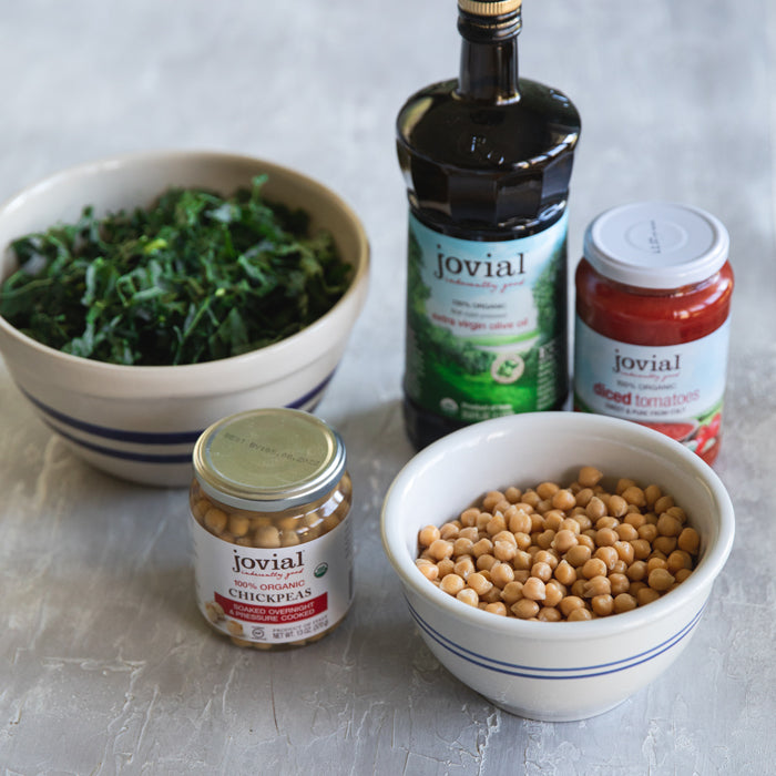 Ingredients for kale chickpea tomato soup arranged on a table with Jovial jars and a bottle of olive oil