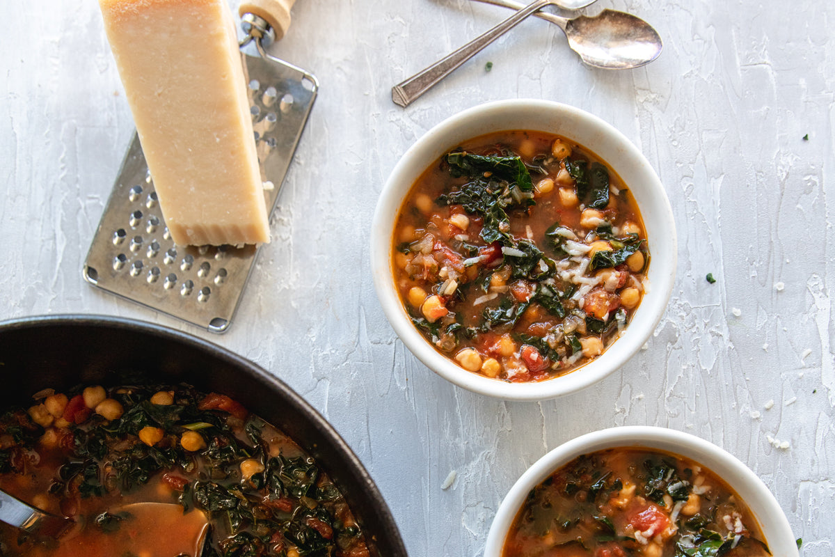 Two bowls of kale and chickpea tomato soup served on a textured white surface with a pot, Parmesan wedge, grater, and spoons