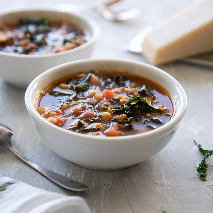 Steaming white bowl of tomato soup with chickpeas and wilted kale, spoon and Parmesan wedge nearby