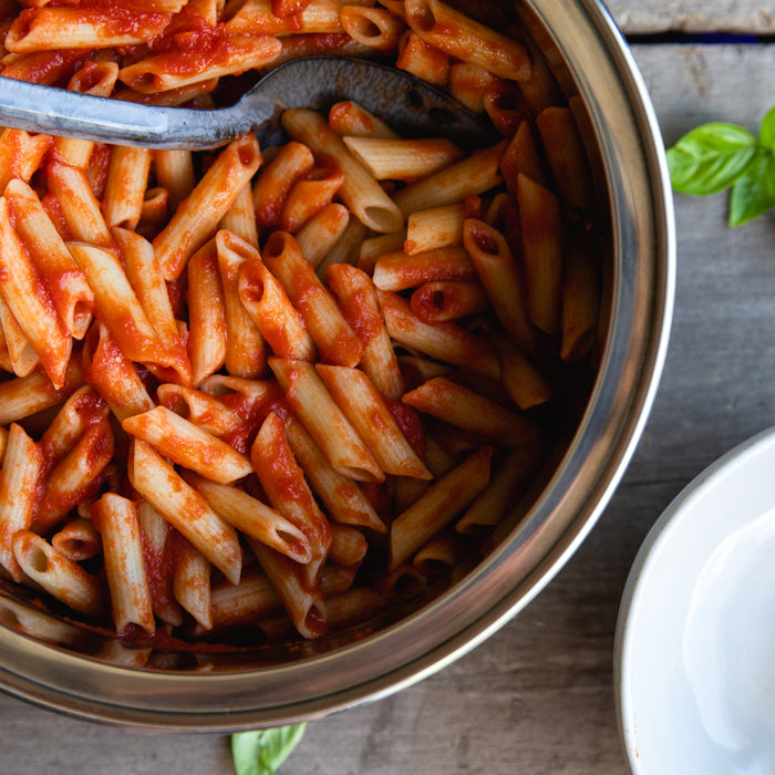 Cooked organic gluten-free rice and lentil penne in tomato sauce in a serving pan with a spoon and basil garnish
