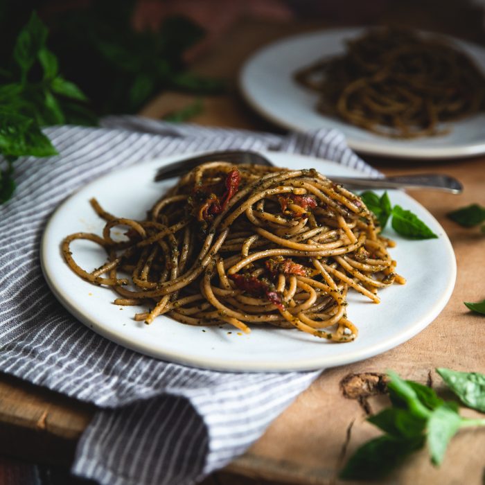 Plate of whole wheat spaghetti tossed with sundried tomato pesto and basil on a white plate with a striped napkin