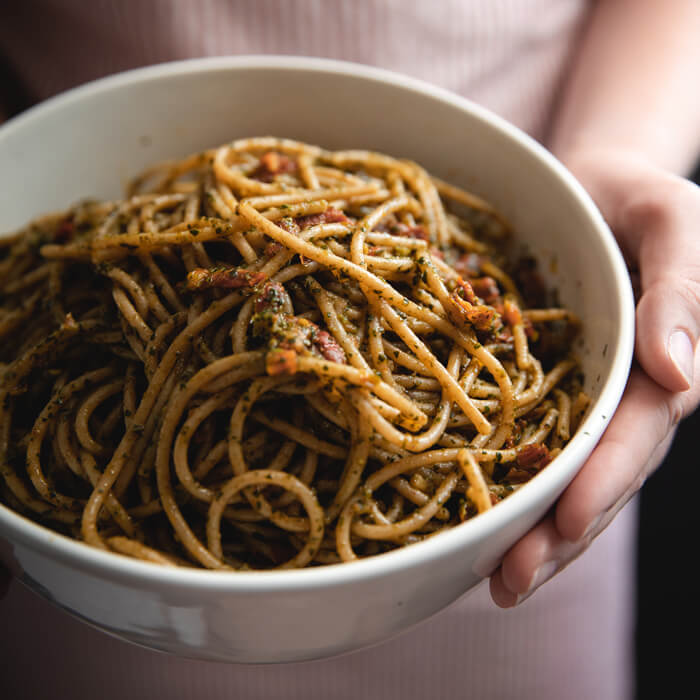 Bowl of organic whole wheat spaghetti tossed with sundried tomato pesto in a white bowl on a wooden table