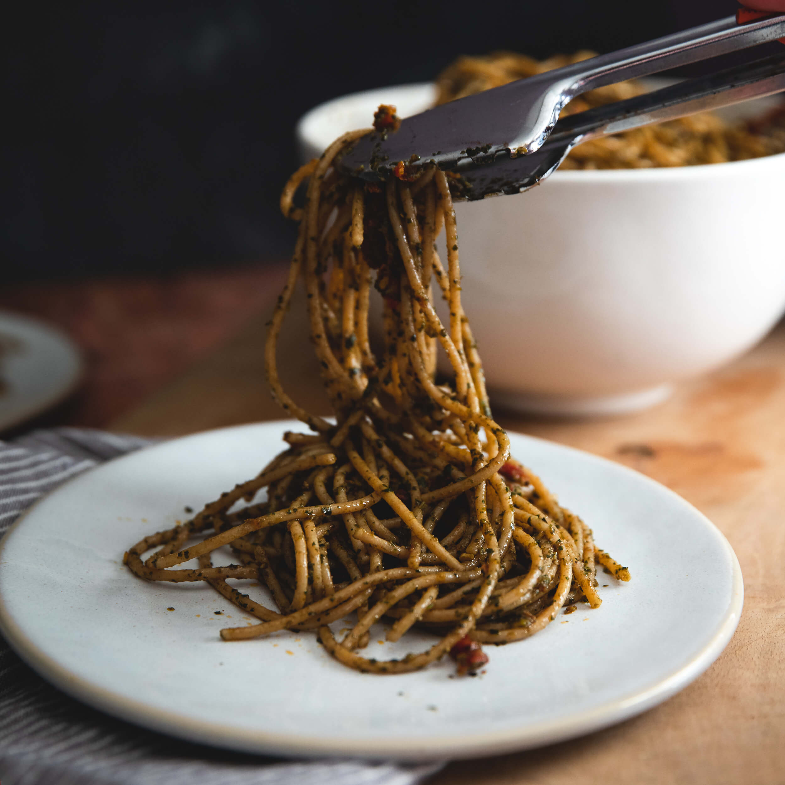 Cooked organic whole wheat spaghetti tossed in sun-dried tomato pesto on a white plate with tongs