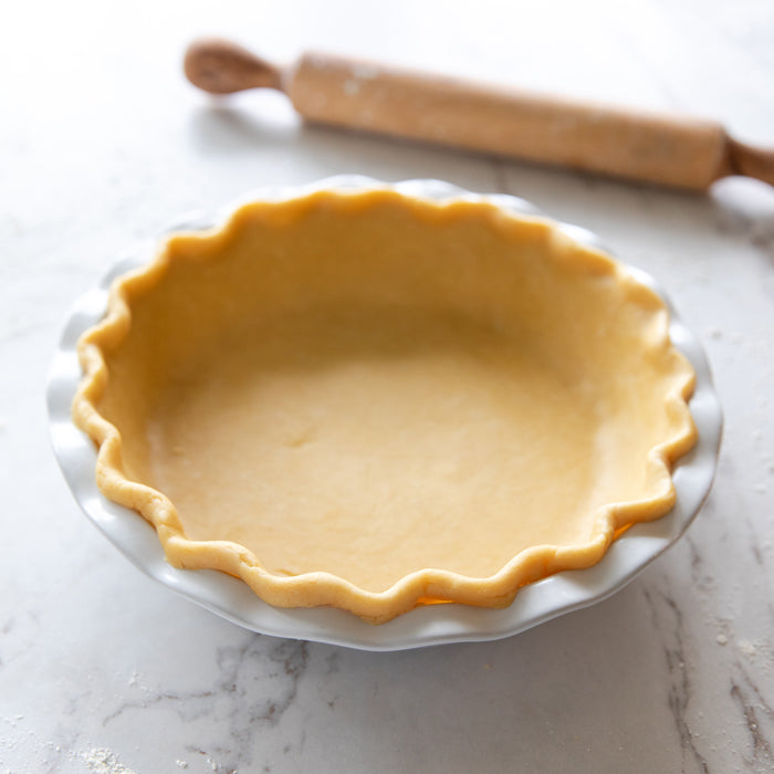 Unbaked einkorn pie crust pressed into a fluted pie dish on a marble countertop with a floured rolling pin in the background