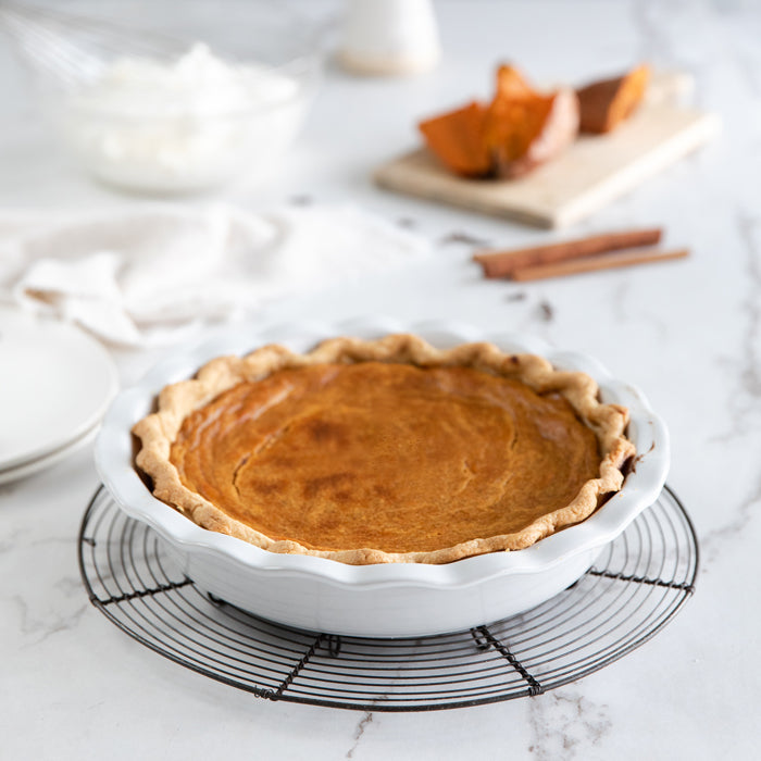 Sweet potato pie with einkorn crust in white pie dish on a cooling rack, sweet potato halves and cinnamon sticks in soft background