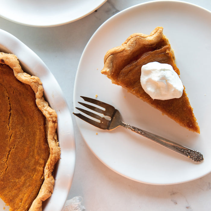 Slice of sweet potato pie with einkorn crust on a white plate topped with whipped cream and a fork beside the remaining pie