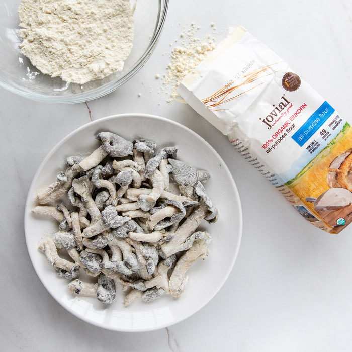 Top-down of einkorn-flour coated mushrooms in a bowl with a Jovial einkorn flour bag and small bowl of flour on marble