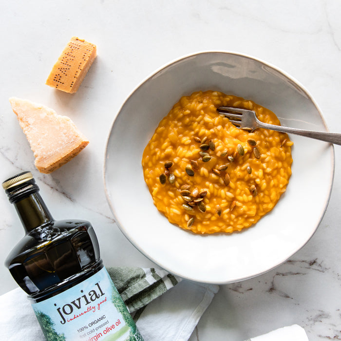 Butternut squash risotto with toasted pepitas, Parmesan wedge, fork, and a bottle of Jovial olive oil on a marble surface