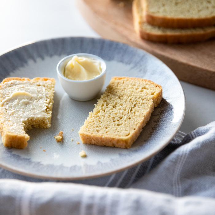 Two slices of einkorn bread on a plate, one spread with butter, dish of butter beside and loaf slices on a wooden board
