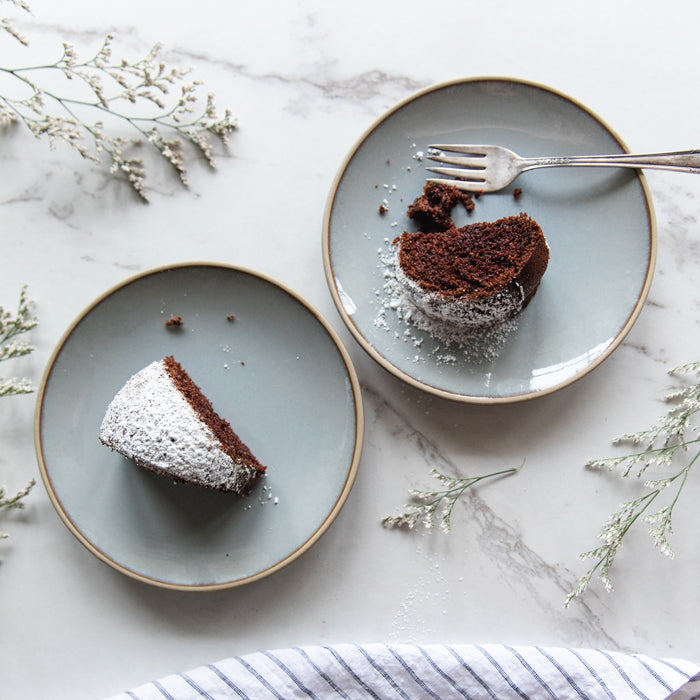 Two slices of dairy-free chocolate olive oil Bundt cake on gray plates, one partially eaten with fork and powdered sugar