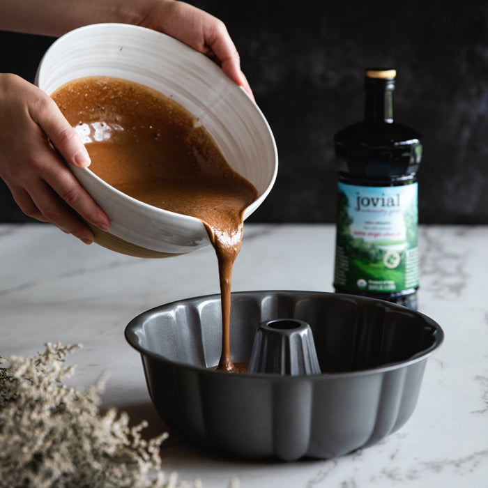 Hands pouring chocolate bundt cake batter from a bowl into a Bundt pan with a bottle of Jovial olive oil in the background