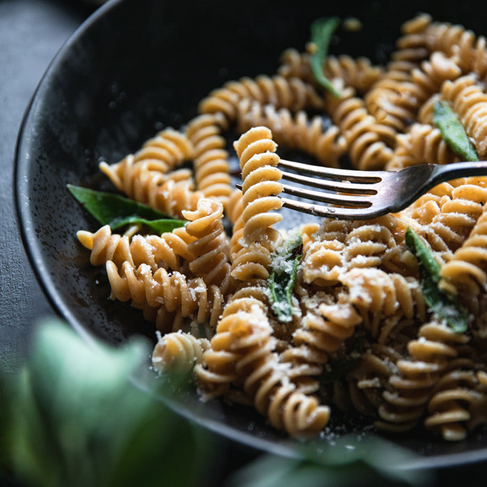 Organic whole wheat fusilli with garlic, sage, and grated Parmigiano in a black bowl, fork lifting one spiral