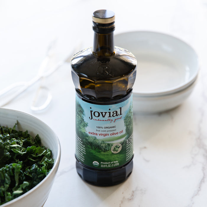 Front view of Jovial organic extra virgin olive oil bottle showing label and dark glass with bowls and chopped kale in background