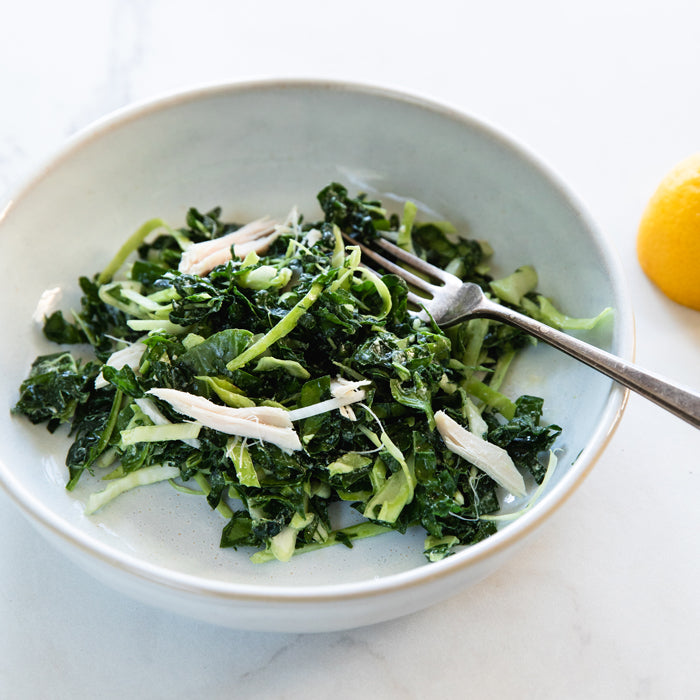Top-down view of winter kale salad with shredded chicken and shaved Brussels sprouts in a white bowl with fork and lemon