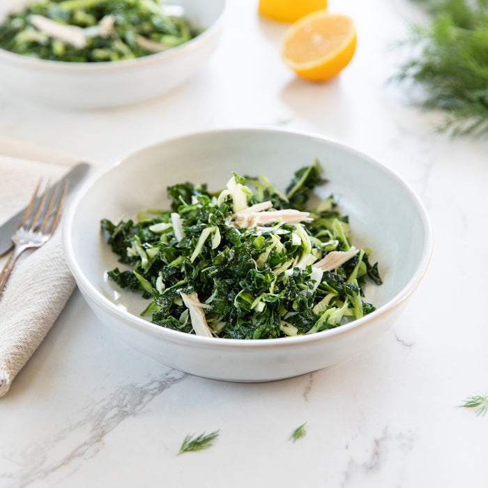 Front-facing bowl of winter kale salad with shredded kale, shaved Brussels sprouts, and pulled chicken on a light marble surface