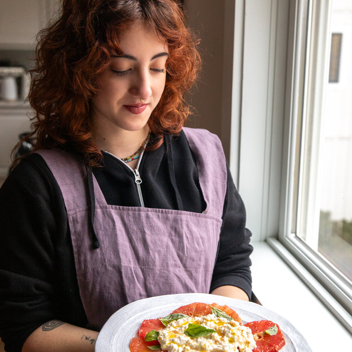 Woman holding a plate of grapefruit and burrata salad garnished with basil and olive oil near a window