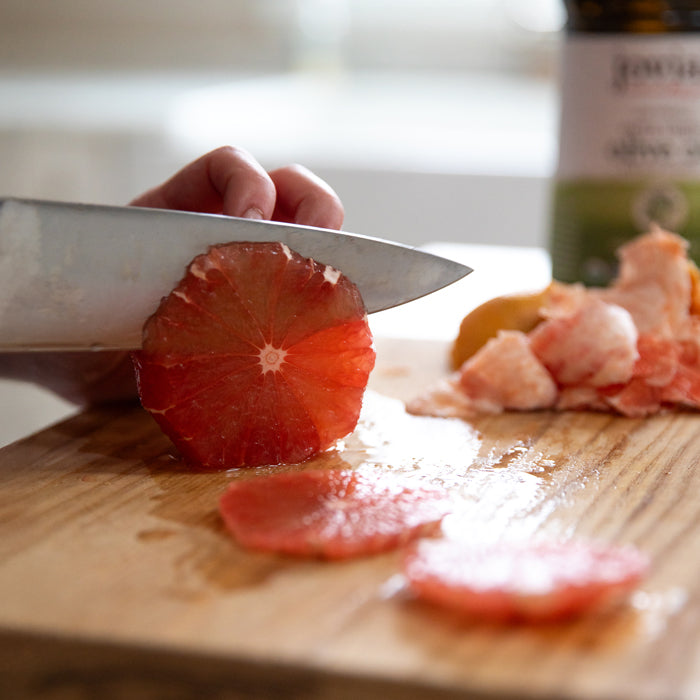 Hand slicing peeled grapefruit into thin rounds on a wooden cutting board with an olive oil bottle blurred in the background