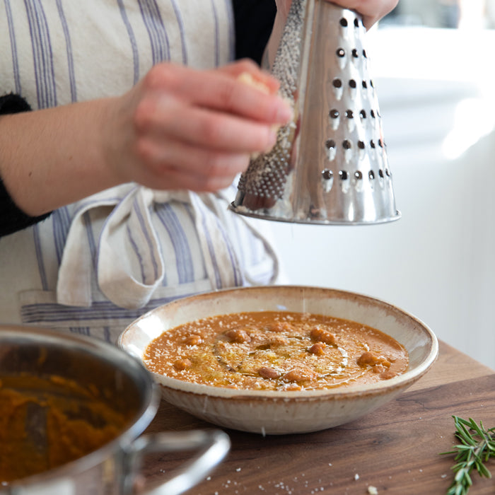 Person grating cheese over a bowl of creamy borlotti bean soup on a wooden counter with a sprig of rosemary