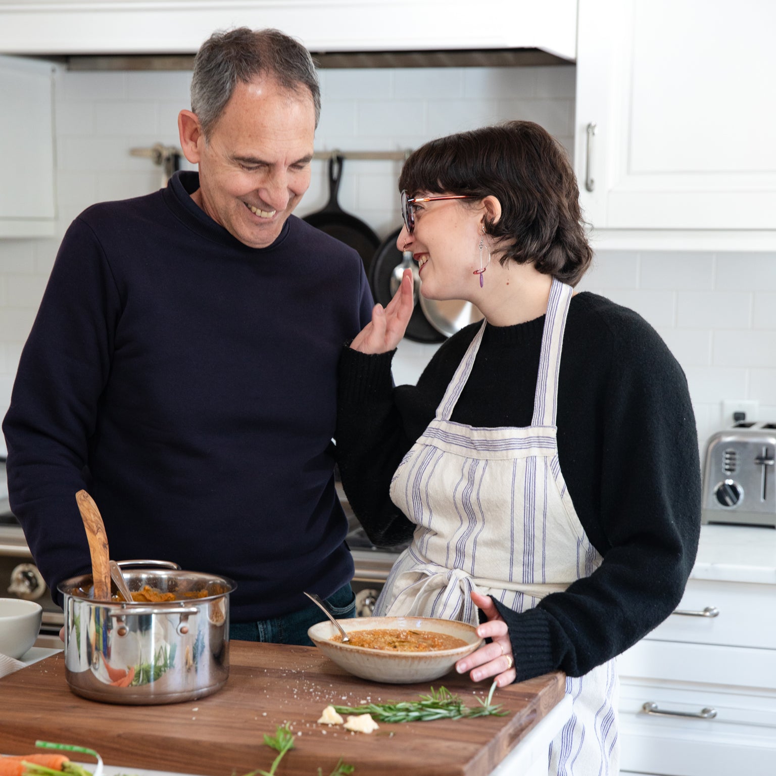 Woman in striped apron tastes a bowl of creamy bean soup while smiling with a man in a bright home kitchen