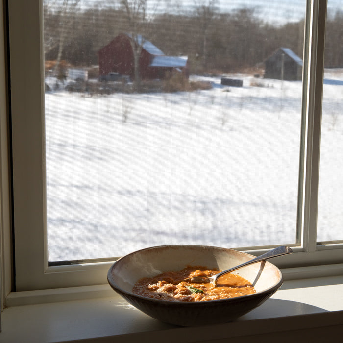 Bowl of creamy Borlotti bean soup on a windowsill with a snow-covered field and red barn outside