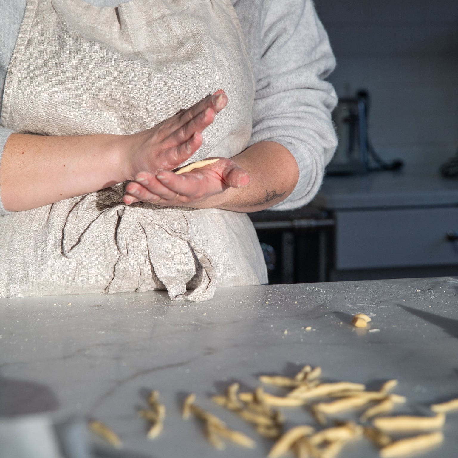Hands shaping trofie-style einkorn pasta dough on a floured marble countertop, person wearing a linen apron in a kitchen