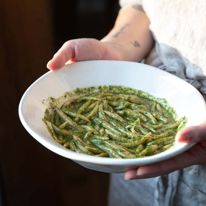 Hands holding a white bowl of homemade einkorn trofie pasta tossed in green pesto, person wearing an apron in the background