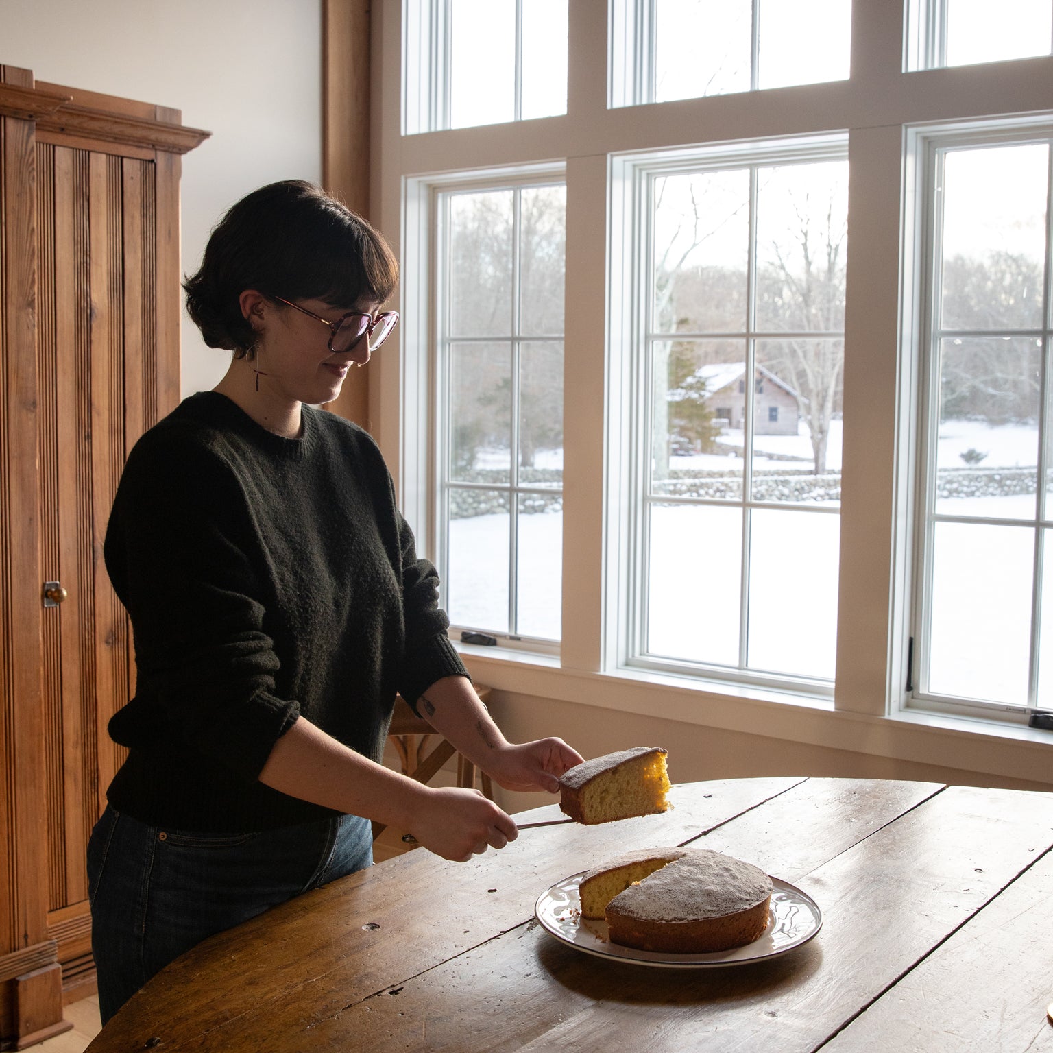 Person slicing an einkorn lemon olive oil cake on a plate at a wooden table by large windows showing a snowy yard