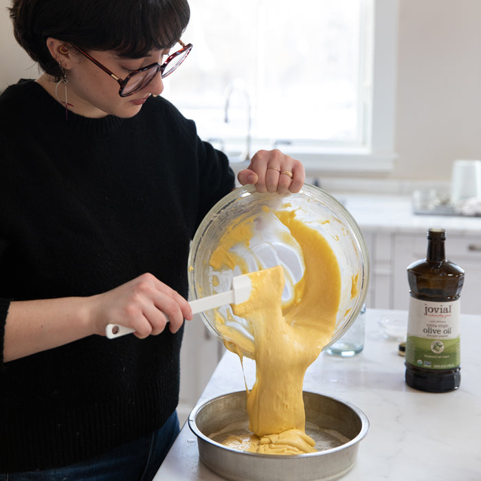 Person pouring einkorn lemon olive oil cake batter into a round pan, Jovial olive oil bottle on the counter
