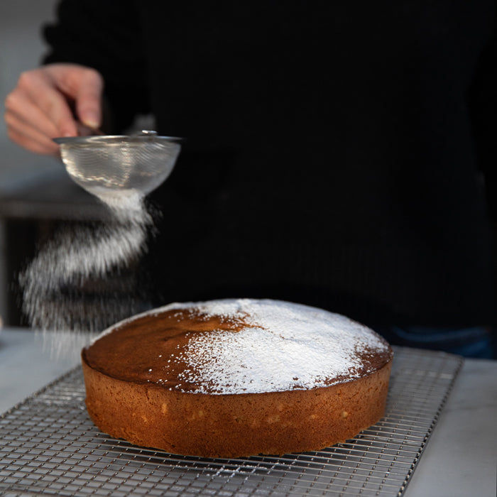 Einkorn lemon olive oil cake being dusted with powdered sugar on a cooling rack, hand holding a sieve in a home kitchen
