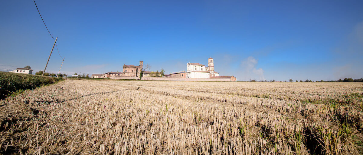 Harvested brown rice field with traditional Italian farm buildings and a white church under a clear blue sky