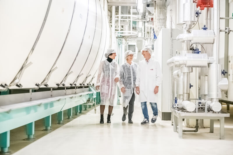 Three people in hairnets and coats walking through an artisan pasta manufacturing facility with large stainless-steel machines
