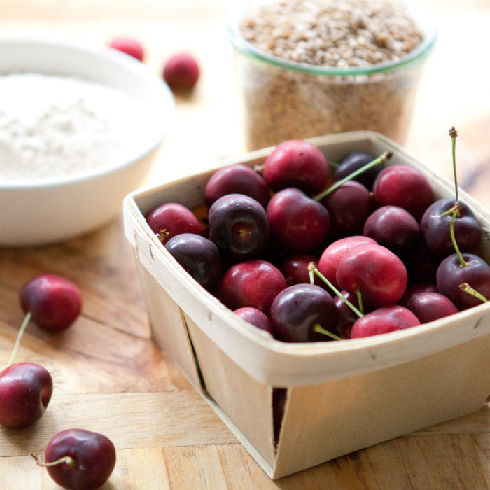 Basket of fresh cherries on a wooden cutting board with bowls of flour and einkorn wheat berries in the background