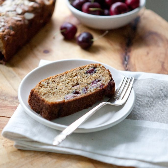 Angled slice of whole-grain einkorn cherry almond quick bread showcasing cherries and sliced almonds on a white plate with wooden table background