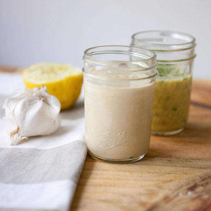 Two mason jars of vinaigrette, creamy garlic and herb versions, on a wooden board with garlic and lemon