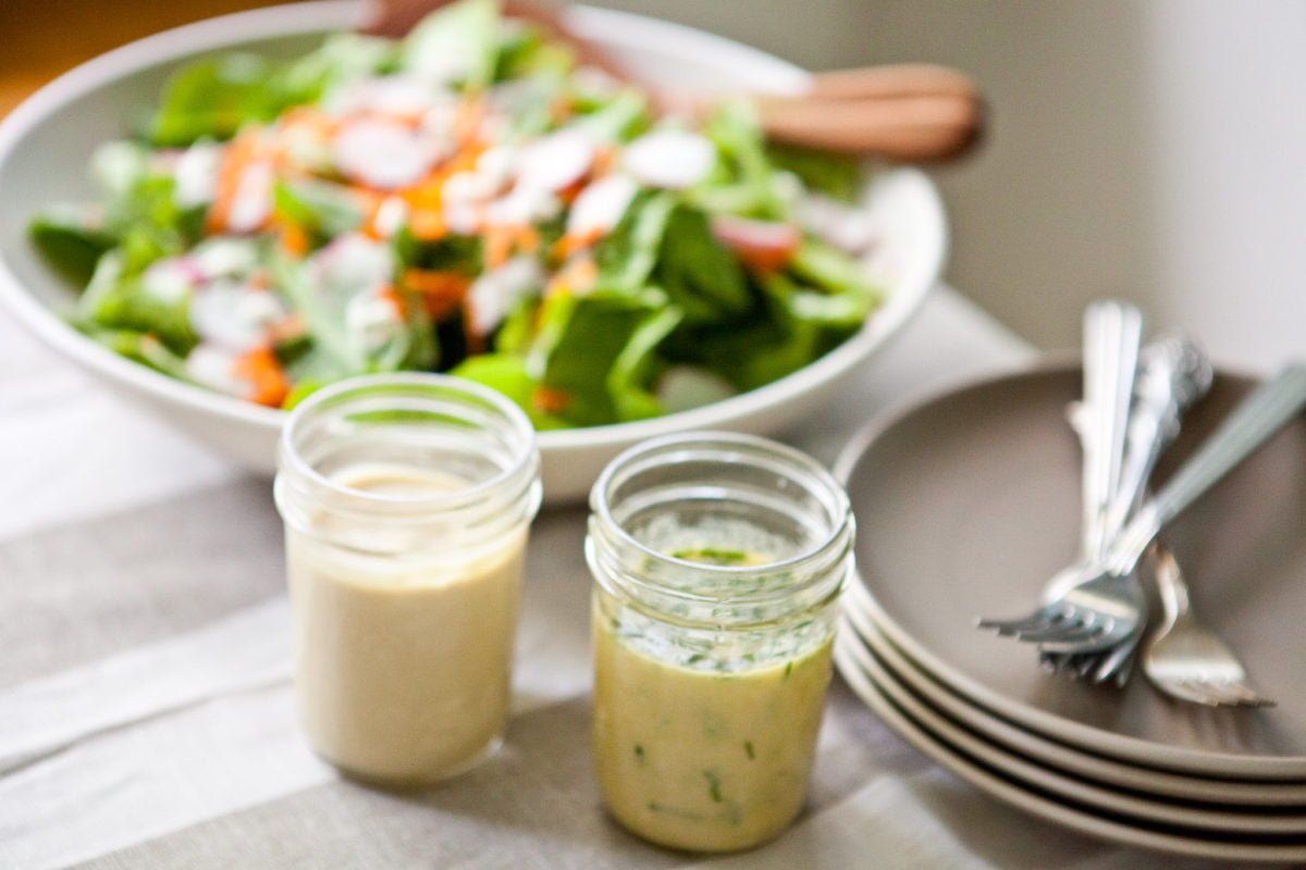 Two small jars of organic garlic herb vinaigrette sit on a table in front of a bowl of mixed green salad and stacked plates