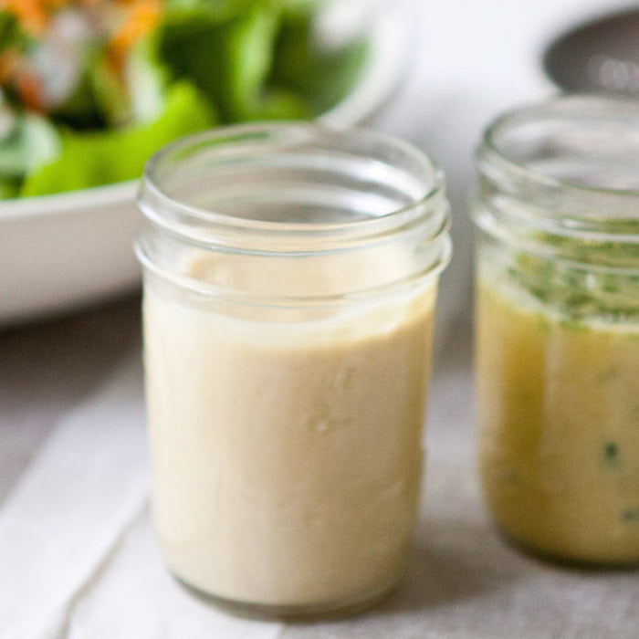 Close-up of two glass jars of vinaigrette, one creamy organic garlic-herb dressing and one oil-based herb vinaigrette, salad blurred behind