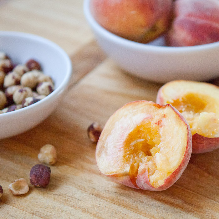 Halved ripe peach on a wooden board with toasted hazelnuts and a bowl of whole peaches in the background