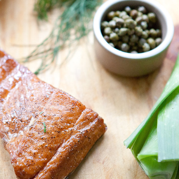 Smoked salmon fillet resting on a wooden board with a bowl of capers, leeks, and dill, ingredients for a salmon tart
