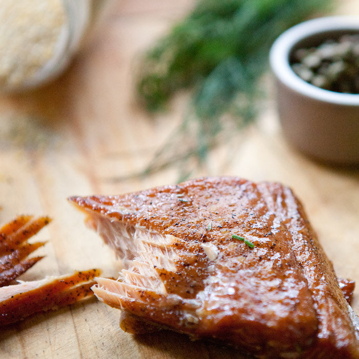 Close-up of smoked salmon showing flaky texture and seasoning on a wooden board with herbs and capers in background